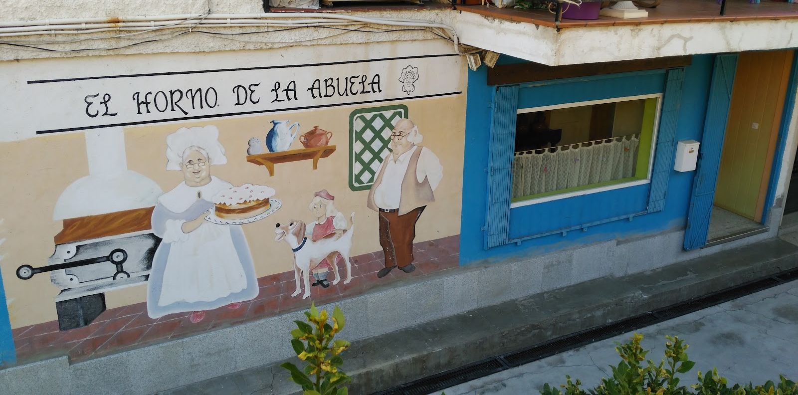 Fachada de El Horno de la Abuela con su mural pintado: una abuela con delantal sacando una tarta del horno junto al abuelo, una niña y un perro, y la ventana azul de la tienda